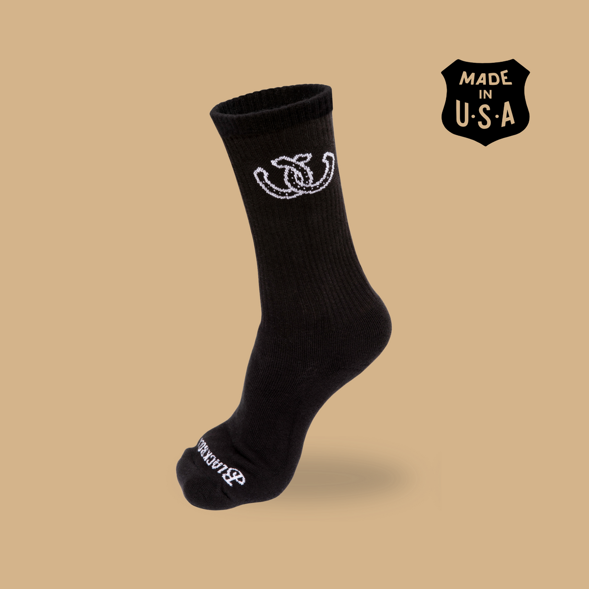 A single black crew sock with a white embroidered interlocking logo near the cuff and white text near the toe, shown floating on a warm beige background with a black 'MADE IN U.S.